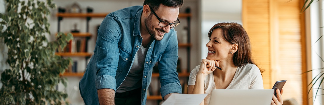 A smiling couple reviews a document; there is a laptop before them on a table