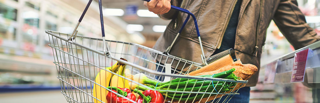 Person holding a shopping basket filled with organic produce in a grocery store aisle.