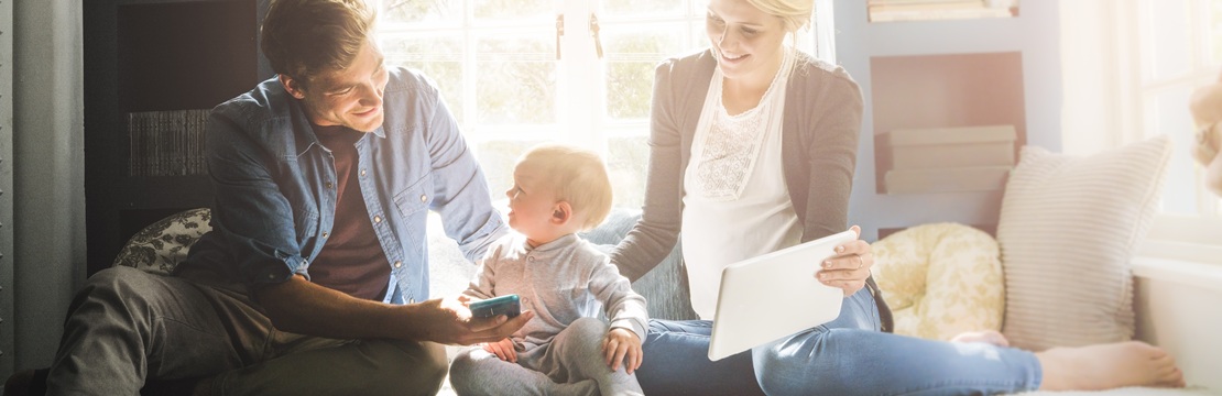 A young family sits together by a bright window, using a tablet and smartphone while discussing tax planning.